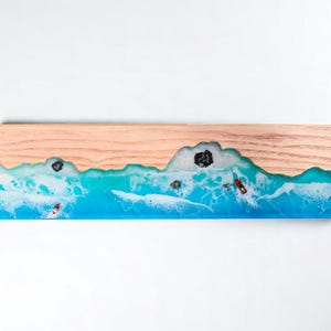 Puede incluir: Tabla de servir rectangular de madera con una escena de playa. La parte superior es de madera marrón claro, que transiciona a un diseño de océano turquesa y blanco con pequeños kayaks rojos. Rocas negras incrustadas en el océano.