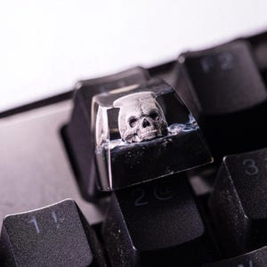 May include: A custom keyboard keycap featuring a detailed, gray skull encased in clear resin. The keycap is black and sits among other black keys on a keyboard. The skull is the focal point, adding a unique, gothic aesthetic.