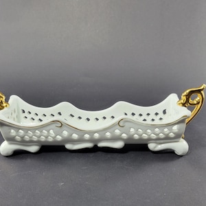 Puede incluir: Un plato de porcelana blanco con un diseño calado y detalles dorados. El plato tiene un borde superior ondulado y asas doradas. Se apoya sobre pequeños pies curvos.