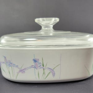 May include: White oval casserole dish with a clear glass lid. The dish features a floral design of purple and blue irises with green stems. The lid has a clear glass knob handle.