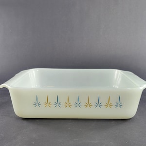 May include: A rectangular white milk glass baking dish with integrated side handles. The exterior features a repeating horizontal pattern of alternating blue and gold starburst or leaf-like motifs arranged in a single row along the front side.