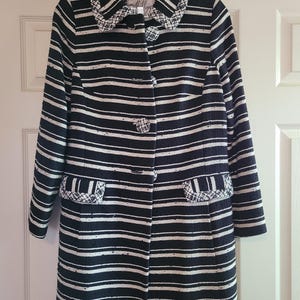 May include: A black and white striped coat with a collared neckline and two front pockets. The coat has a button-up closure and is hanging on a wooden hanger. The coat's design features horizontal stripes.
