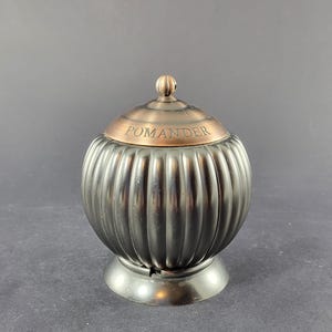 Peut inclure: Un pot-pourri gris foncé et nervuré avec un couvercle cuivré et un petit bouton sur le dessus. Le mot "POMANDER" est gravé sur le couvercle. Le pot-pourri repose sur une petite base ronde.