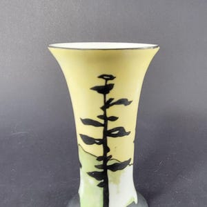 May include: A small, decorative vase with a flared top and a black base. The vase has a pale yellow background with a black tree design. The tree design is stylized and appears to be a silhouette of a pine tree. The vase is approximately 6 inches tall.
