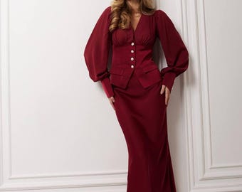 May include: A burgundy two-piece suit featuring a fitted blazer with gold buttons and a matching midi skirt. The blazer has long, voluminous sleeves. The outfit is paired with black pointed-toe heels. The background is a white wall with decorative molding.