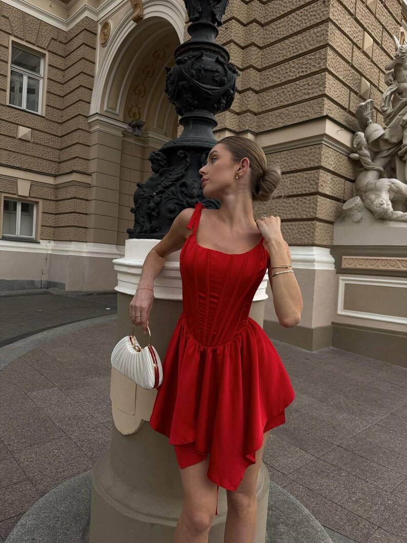 Puede incluir: Un vestido rojo vibrante con un corpi&ntilde;o estilo cors&eacute; y una falda acampanada. El vestido presenta delicadas correas para los hombros y un dobladillo asim&eacute;trico &uacute;nico. Se sostiene en la mano un bolso de mano blanco y beige con asa dorada.