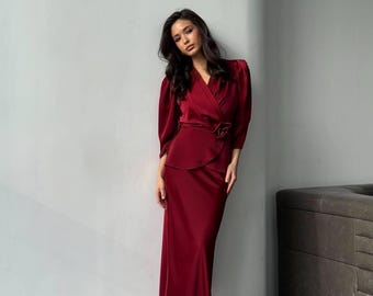 May include: A woman wearing a long, burgundy satin dress with a wrap-style bodice and a matching belt. The dress has a slightly flared skirt and the woman is wearing white heels.