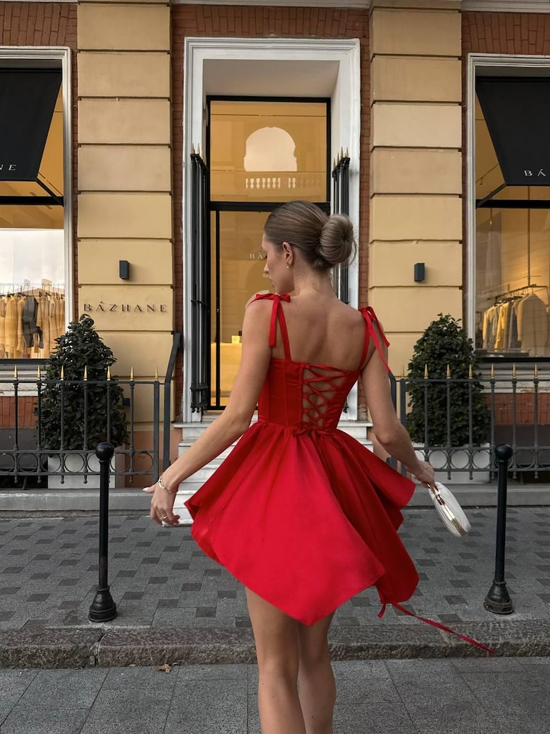 Puede incluir: Un vestido rojo vibrante con espalda estilo cors&eacute; y falda acampanada. El vestido presenta tirantes anudados y un dise&ntilde;o fluido. La imagen muestra el vestido en movimiento, sugiriendo una tela ligera. El fondo incluye un edificio con el texto "BAZHANE".