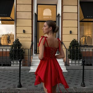 Puede incluir: Un vestido rojo vibrante con un dobladillo asim&eacute;trico &uacute;nico y una espalda con cordones. El vestido presenta delicadas correas anudadas y una falda fluida. El fondo incluye una fachada de edificio con el nombre "BAZHANE" visible en el toldo.