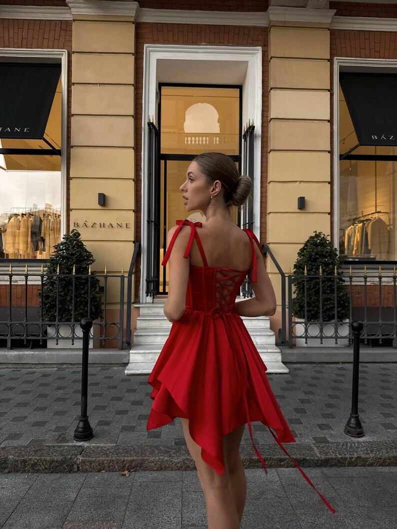Puede incluir: Un vestido rojo vibrante con un dobladillo asim&eacute;trico &uacute;nico y una espalda con cordones. El vestido presenta delicadas correas anudadas y una falda fluida. El fondo incluye una fachada de edificio con el nombre "BAZHANE" visible en el toldo.