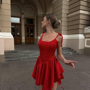 Puede incluir: Un vestido rojo vibrante con un corpi&ntilde;o estilo cors&eacute; y una falda acampanada de varias capas. El vestido presenta delicadas correas anudadas y una silueta favorecedora. El vestido est&aacute; hecho de una tela suave y fluida, ideal para ocasiones especiales.