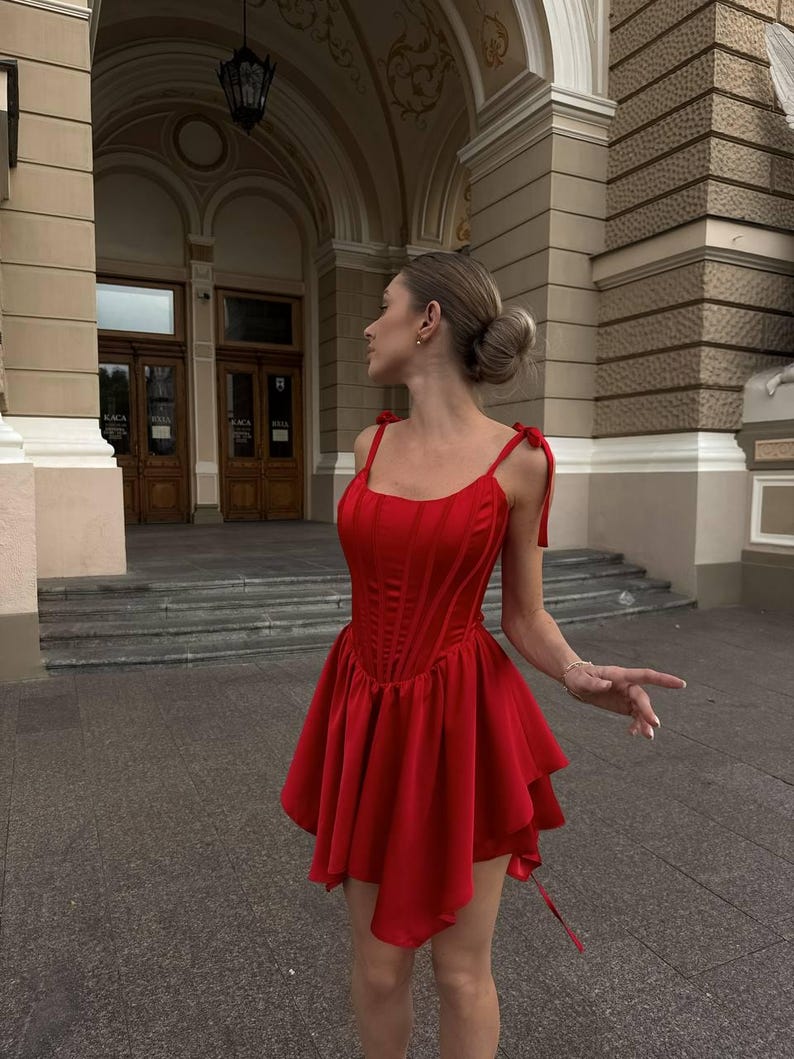 Puede incluir: Un vestido rojo vibrante con un corpi&ntilde;o estilo cors&eacute; y una falda acampanada de varias capas. El vestido presenta delicadas correas anudadas y una silueta favorecedora. El vestido est&aacute; hecho de una tela suave y fluida, ideal para ocasiones especiales.