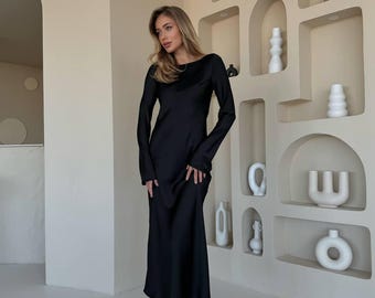 May include: A woman wearing a black long-sleeved maxi dress with a cowl neck. The dress is made of a silky fabric and has a flowing silhouette. She is standing in front of a wall with decorative niches.