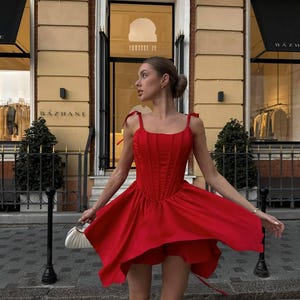 Puede incluir: Un vestido rojo vibrante con un corpi&ntilde;o estilo cors&eacute; y una falda fluida. El vestido presenta delicadas correas anudadas y una cintura ce&ntilde;ida. La imagen est&aacute; tomada al aire libre, mostrando el movimiento y el dise&ntilde;o del vestido. El vestido se combina con un peque&ntilde;o bolso blanco.