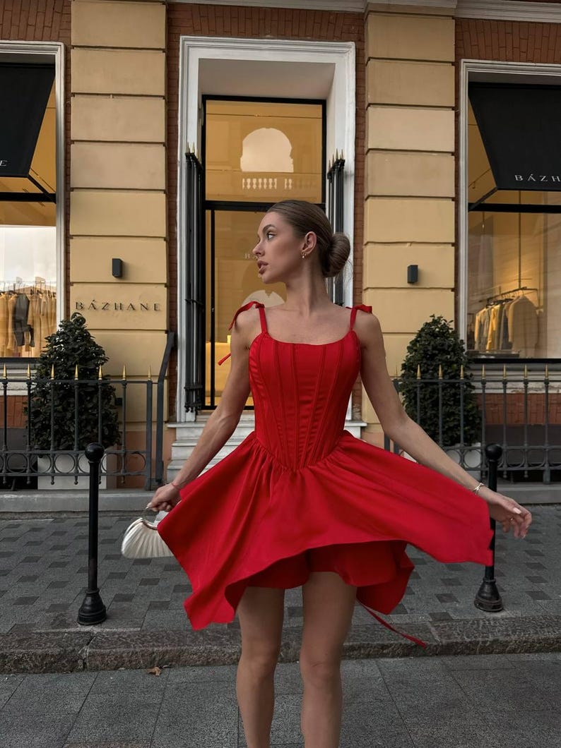 Puede incluir: Un vestido rojo vibrante con un corpi&ntilde;o estilo cors&eacute; y una falda fluida. El vestido presenta delicadas correas anudadas y una cintura ce&ntilde;ida. La imagen est&aacute; tomada al aire libre, mostrando el movimiento y el dise&ntilde;o del vestido. El vestido se combina con un peque&ntilde;o bolso blanco.