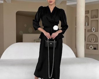 May include: A woman wearing a black satin wrap top with a white flower accent and a matching black satin skirt. She is holding a small black quilted handbag with a silver chain.