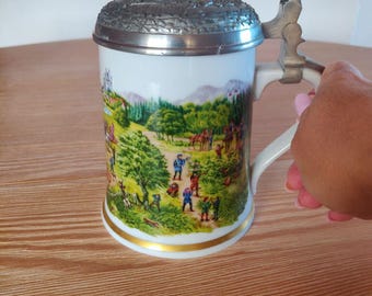 Könnte beinhalten: Ein weißer Bierkrug aus Keramik mit einer detaillierten Landschaftsszene mit Figuren, Bäumen und Pferden. Der Krug hat einen Metalldeckel und einen Griff sowie ein goldenes Band am Boden.