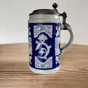 Op de afbeelding: Een blauw-witte keramische bierpul met een scharnierend deksel. De pul heeft een gedetailleerd bloemen- en dierenontwerp. De handgreep en het deksel zijn een gedempte grijze kleur. De pul staat op een houten oppervlak.
