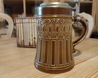 May include: A brown ceramic beer stein with a decorative pattern and a metal hinged lid. The stein has a large handle and is sitting on a wooden surface. The stein is a traditional style and is likely used for drinking beer.