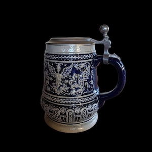 Può includere: Un boccale da birra in ceramica blu e bianca con coperchio e manico in metallo. Il corpo presenta intricati motivi in rilievo, tra cui una figura alata centrale circondata da motivi floreali e un bordo geometrico alla base e sul bordo superiore.