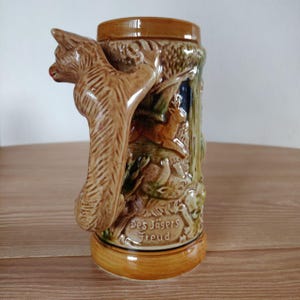 Pode incluir: Um caneco de cerveja de cerâmica com um design de relevo detalhado. A alça tem a forma de um esquilo. O caneco é castanho e bege com uma cena de floresta e as palavras "Des Jägers Freud" em uma fonte decorativa.