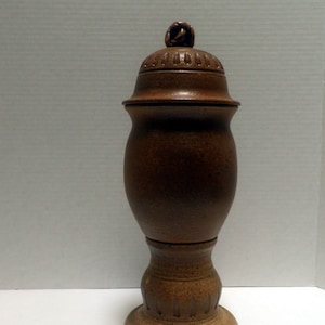 May include: A brown ceramic jar with a lid and a pedestal base. The jar has a textured surface and a decorative design around the top and bottom.