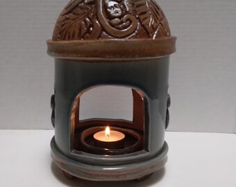 May include: A ceramic candle holder with a domed top featuring a brown floral design. The holder is a light blue colour and has a small opening for a candle. A small white candle is burning inside the holder.