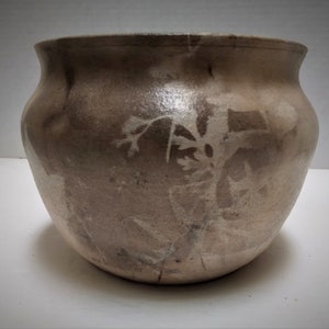 May include: A light brown ceramic pot with a rounded shape and a subtle, mottled pattern. The pot has a smooth, glazed finish.