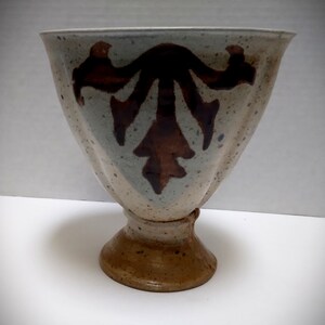 May include: A ceramic vase with a wide, fluted top and a pedestal base. The vase features a speckled, light-colored exterior with a dark brown, stylized leaf design. The base is a darker brown, creating a contrast with the upper portion.