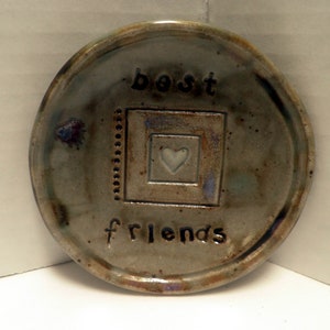 May include: A ceramic coaster with a gray glaze and a heart design. The coaster has the words "best friends" written on it.