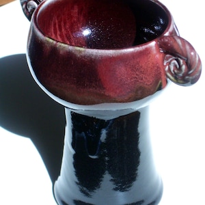 May include: A red and black ceramic vase with two handles shaped like rams' horns. The vase has a wide, round opening and a narrow base.