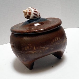 May include: A brown ceramic jar with a lid and three legs. The lid is topped with a white and brown seashell.