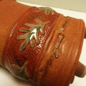 May include: A brown ceramic pot with a decorative design of green and white leaves and branches. The pot has a textured surface and three small feet.