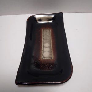 May include: A rectangular, dark brown ceramic dish with a glossy finish. The dish has a unique, asymmetrical shape with a lighter brown and white rectangular design in the center. The dish is likely for decorative or serving purposes.