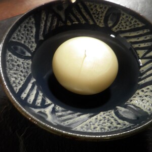 May include: A white candle sits in a blue and white ceramic dish with a decorative pattern. The dish has a dark blue glaze with a white pattern of lines and dots.
