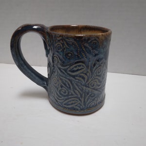 May include: A blue ceramic mug with a brown rim and a swirling, embossed pattern. The mug has a handle.