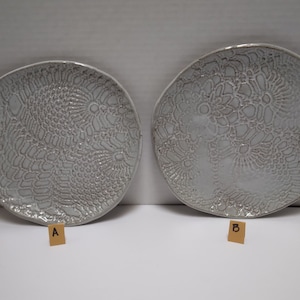 May include: Two white ceramic plates with a raised, lacy design. The plates are labeled A and B.