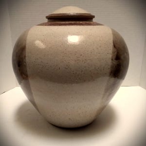 May include: A ceramic urn with a lid. The urn is a light beige color with vertical brown stripes. The lid is a darker brown. The urn has a rounded shape.