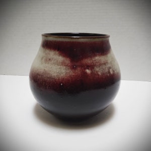 May include: A small, round, ceramic vase with a dark brown glaze and white streaks. The vase has a slightly flared lip and a rounded bottom.
