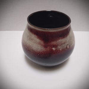 K&ouml;nnte beinhalten: Eine kleine, runde Keramikvase mit einer dunkelbraunen Glasur und einem wei&szlig;en, gesprenkelten Design. Die Vase steht auf einer wei&szlig;en Oberfl&auml;che.