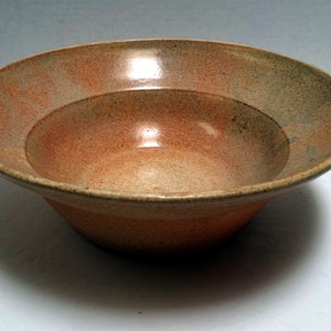 May include: A large, round, ceramic bowl with a light brown glaze and a speckled finish. The bowl has a slightly raised rim and a wide, shallow base.