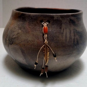 May include: A gray ceramic bowl with a brown and orange design. A white cord with a red bead, a gold feather charm, and black and brown beads hangs from the bowl.