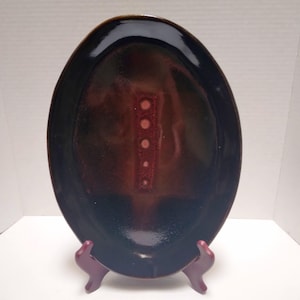 May include: Oval-shaped ceramic serving platter with a glossy finish. The platter features a dark brown exterior that fades into a reddish-brown center with a vertical line of white dots. The platter is displayed on a burgundy stand.