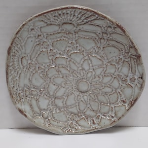 May include: A round, ceramic plate with a detailed, lace-like pattern. The plate is off-white with a brown rim and accents, creating a rustic, handcrafted appearance. The design features a central floral motif, suitable for serving or display.