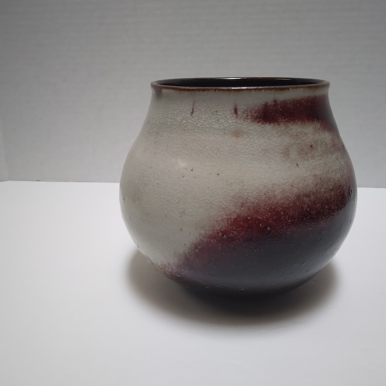 K&ouml;nnte beinhalten: Eine wei&szlig;e Keramikvase mit einem dunkelbraunen Rand und einer burgunderfarbenen Glasur, die in Wei&szlig; &uuml;bergeht. Die Vase hat eine abgerundete, bauchige Form.