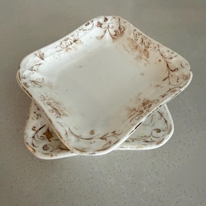 May include: Two white, square-shaped ceramic plates with rounded corners. Each plate features a brown floral pattern around the edges. The plates are stacked on top of each other, showcasing the design.