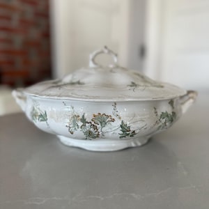 May include: White ceramic serving dish with lid. The dish features a floral pattern in green and gold. The lid has a loop handle. The dish has two side handles. The dish is on a gray surface.