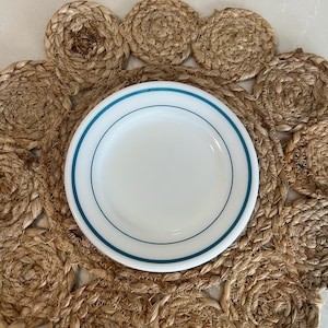 May include: A white plate with a teal blue rim sits on a woven jute placemat. The placemat is circular with a scalloped edge, and the plate is centered on the mat. The natural tones of the jute contrast with the bright white of the plate.