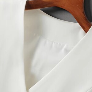 May include: Close-up of a white sleeveless top with a V-neck design. The top is hanging on a wooden hanger. The fabric appears to be smooth and opaque, suggesting a formal or semi-formal garment.