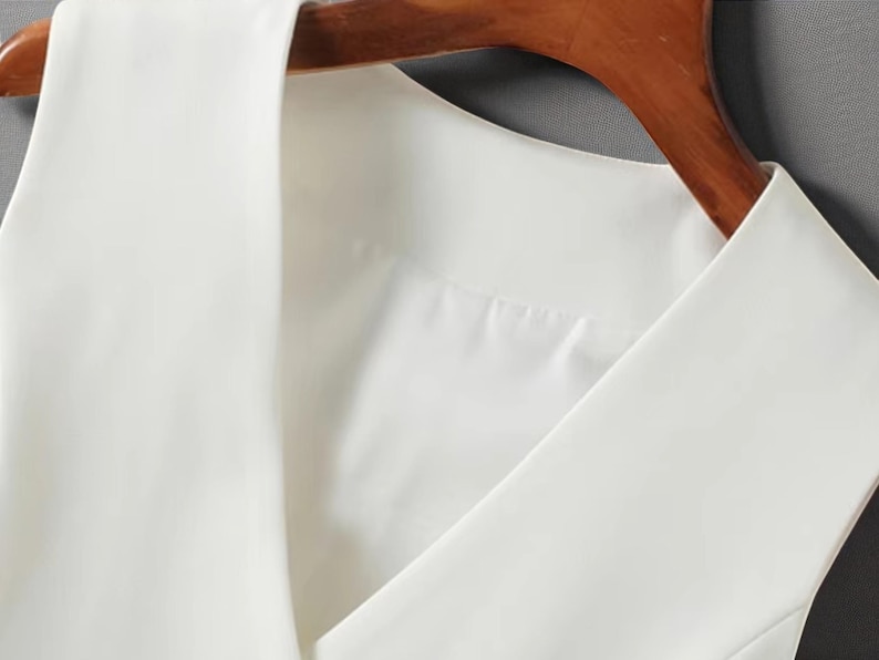 May include: Close-up of a white sleeveless top with a V-neck design. The top is hanging on a wooden hanger. The fabric appears to be smooth and opaque, suggesting a formal or semi-formal garment.
